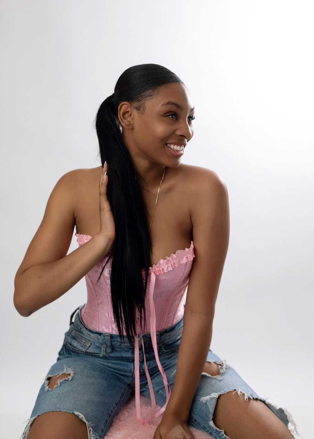 Young woman smiling in pink corset top and ripped denim shorts with long ponytail on gray background.