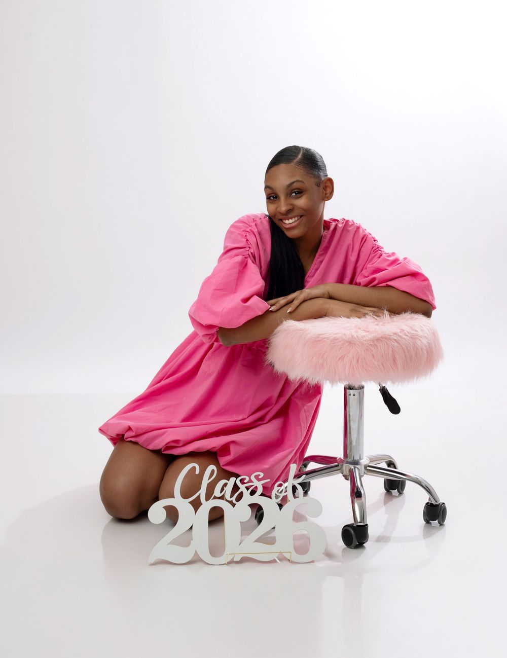 Young woman in pink dress poses with Class of 2025 sign beside a stool for graduation photos.