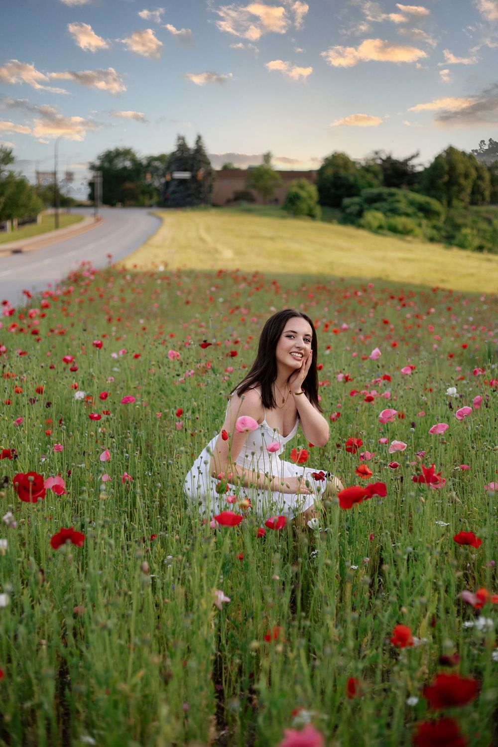 Young woman in white dress crouching among red and pink poppies in a field at sunset.