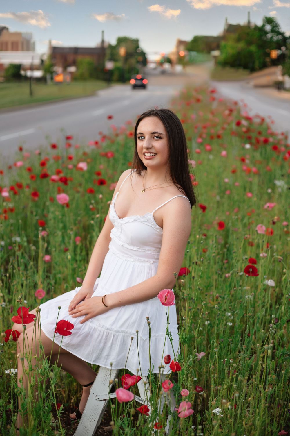 Young woman in white dress sitting among red and pink poppies by a road at sunset.