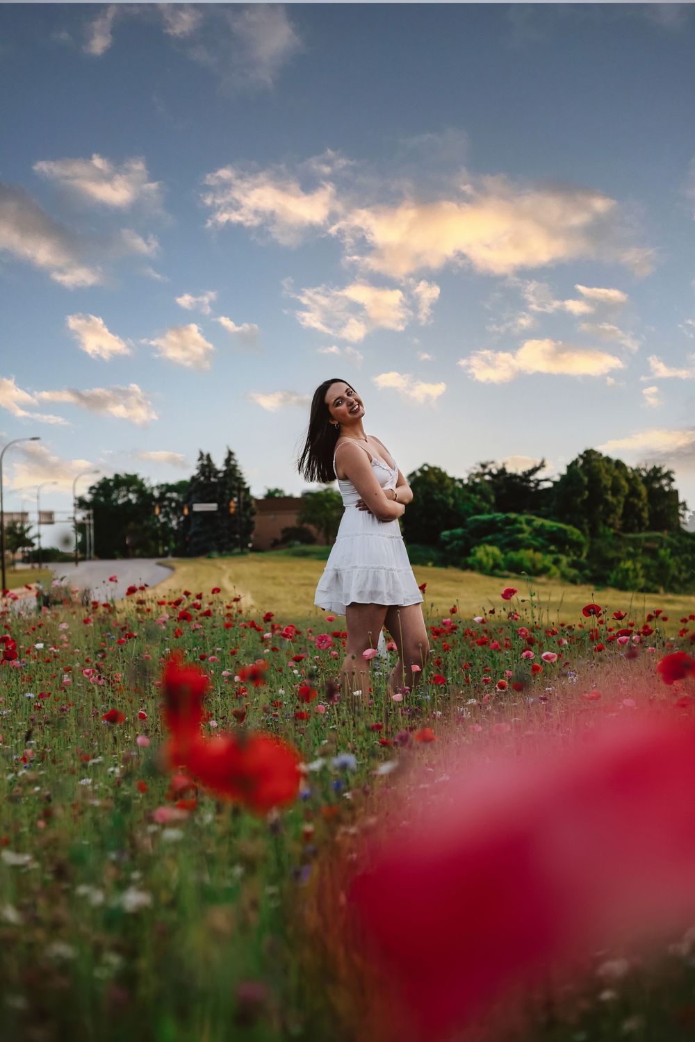 Young woman in white dress standing in vibrant red poppy field at sunset with dramatic cloudy sky.