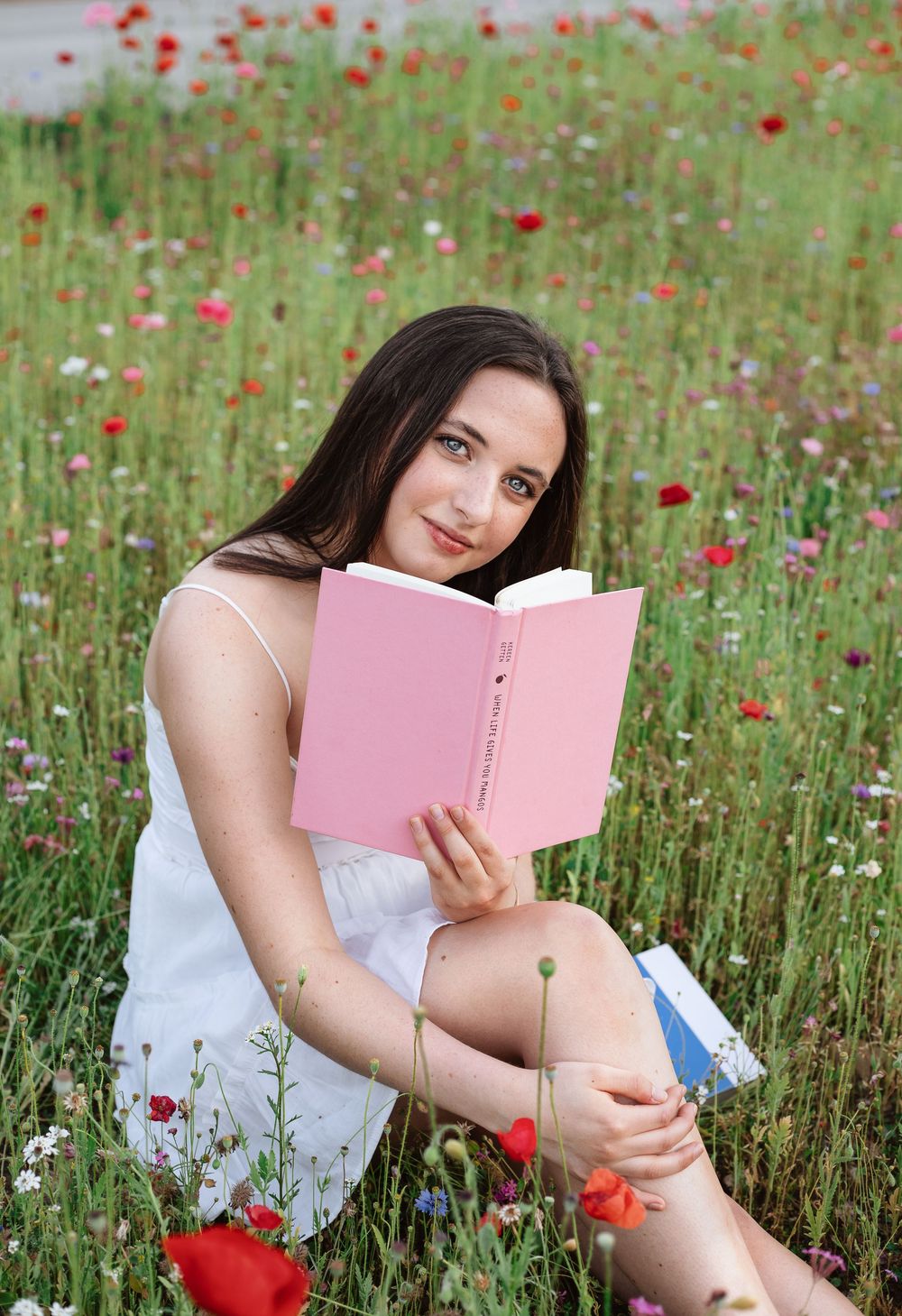 Young woman in white dress reading a pink book while sitting in a vibrant wildflower meadow with red poppies.