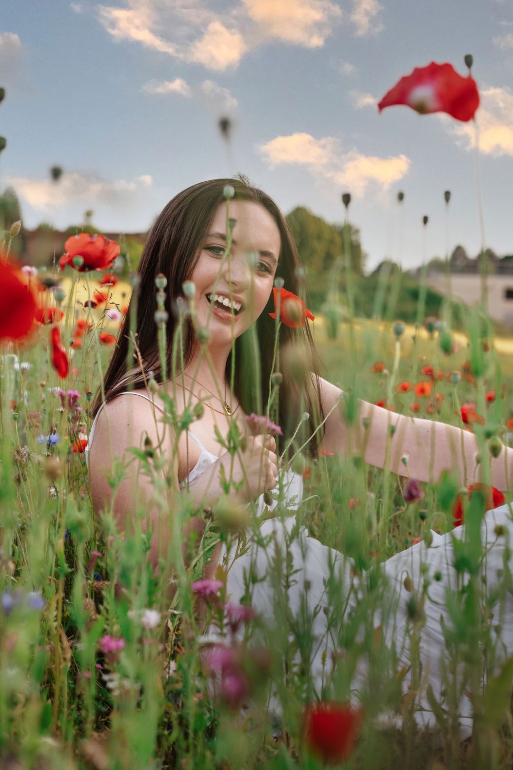 Young woman laughing joyfully while sitting in a vibrant poppy field with wildflowers under a blue sky.