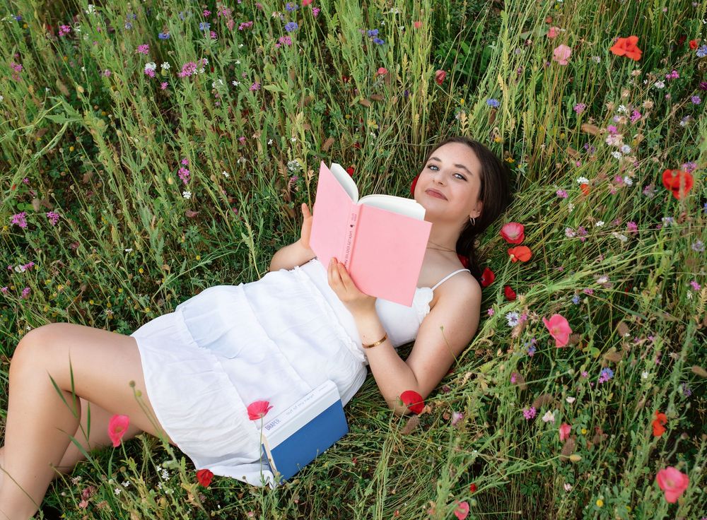 Young woman in white dress lying in a wildflower meadow reading a pink book among red poppies.