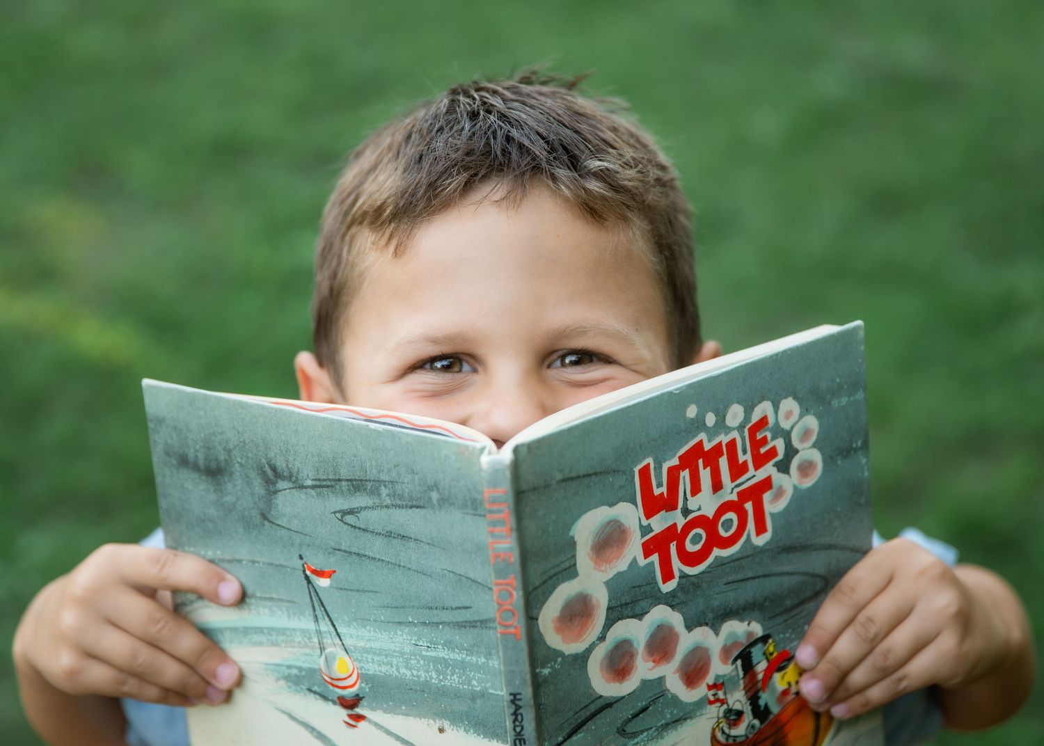 Young boy reading Little Toot children's book outdoors, peeking over the colorful cover with curious eyes.