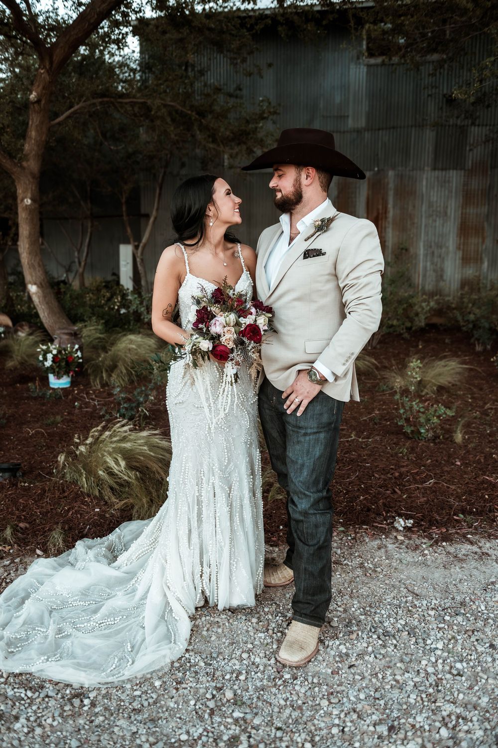 Bride in lace gown holding bouquet stands with groom in cowboy hat and tan blazer at rustic outdoor wedding.