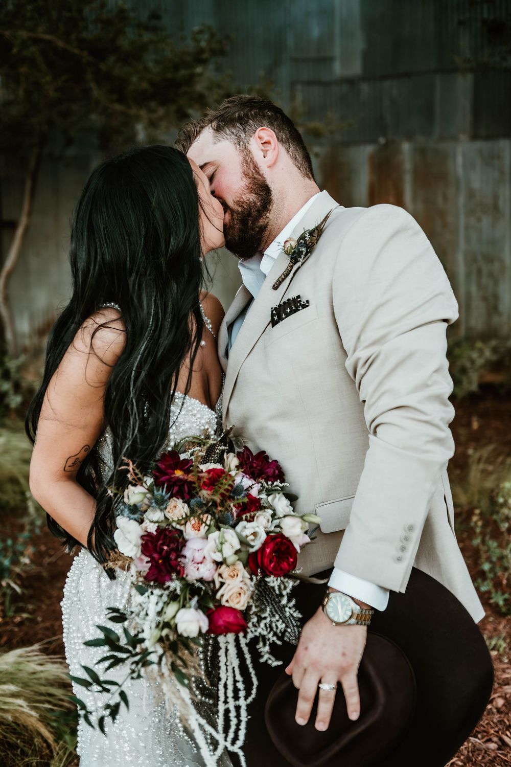 Bride and groom share a romantic kiss, she holds a lush bouquet of red and white flowers, he wears a cream suit.