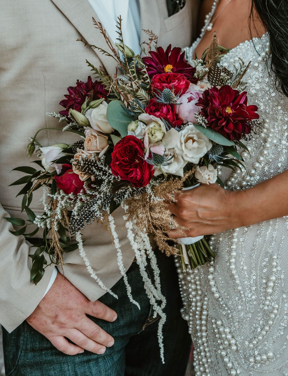 Bride holding a lush wedding bouquet with deep red dahlias, white roses, and wildflowers, standing beside groom in beige suit.