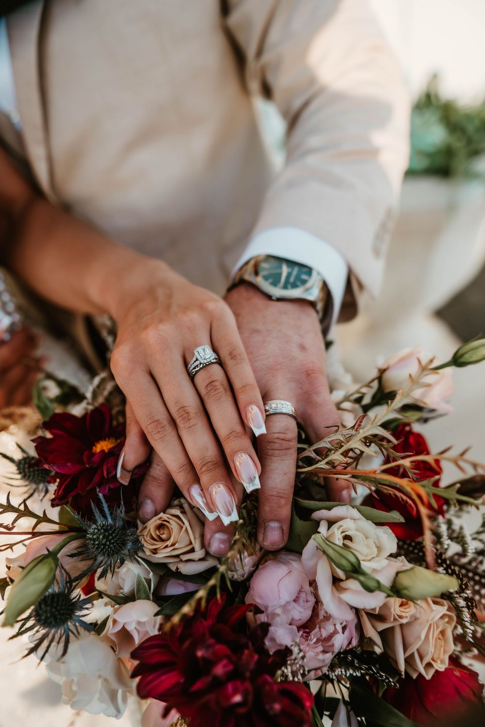 Newlyweds place hands on wedding bouquet showing diamond engagement ring and gold bands with a blue-faced watch.