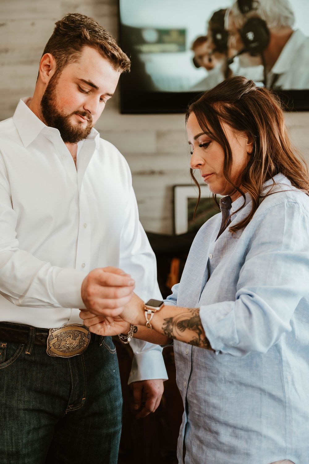 A bearded man in a white shirt and jeans has his watch fastened by a tattooed woman in a blue shirt indoors.