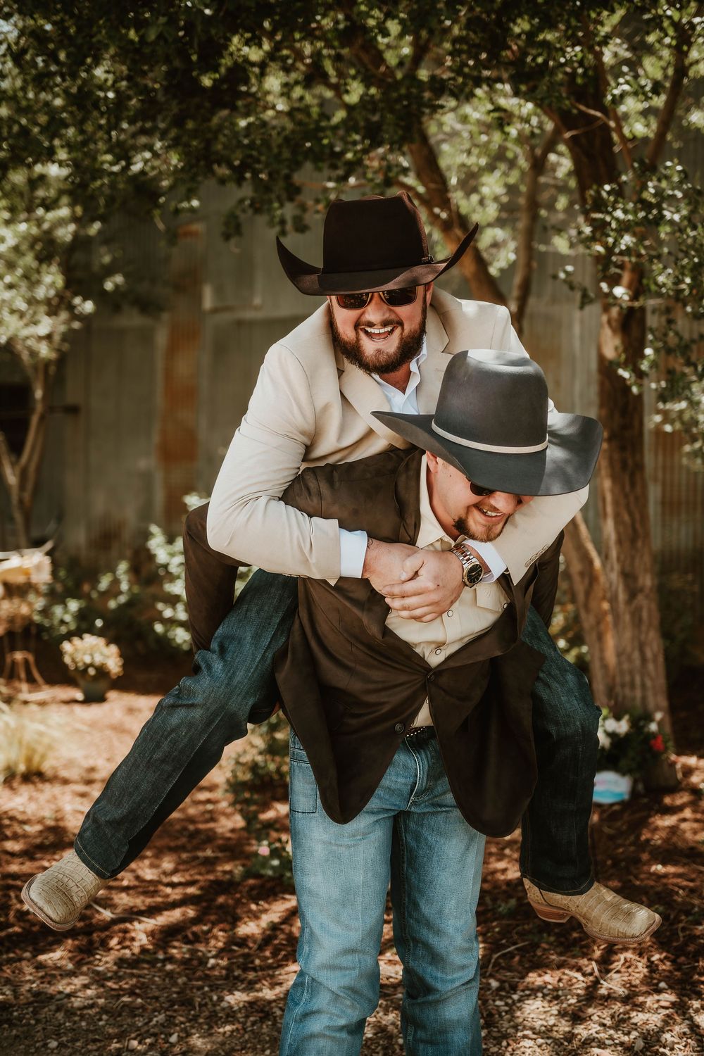 Two men in cowboy hats laugh as one gives the other a piggyback ride outdoors at a rustic wedding venue.