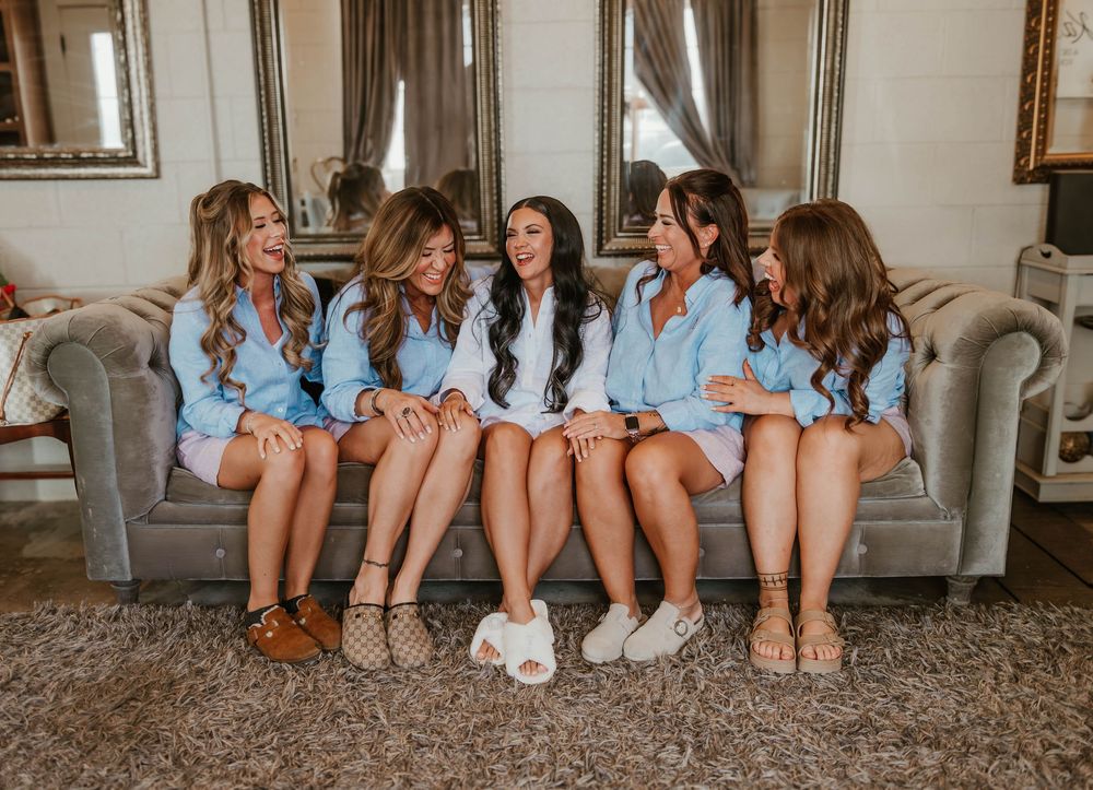 Six bridesmaids in matching light blue robes sitting together on a gray sofa, laughing and celebrating before a wedding.
