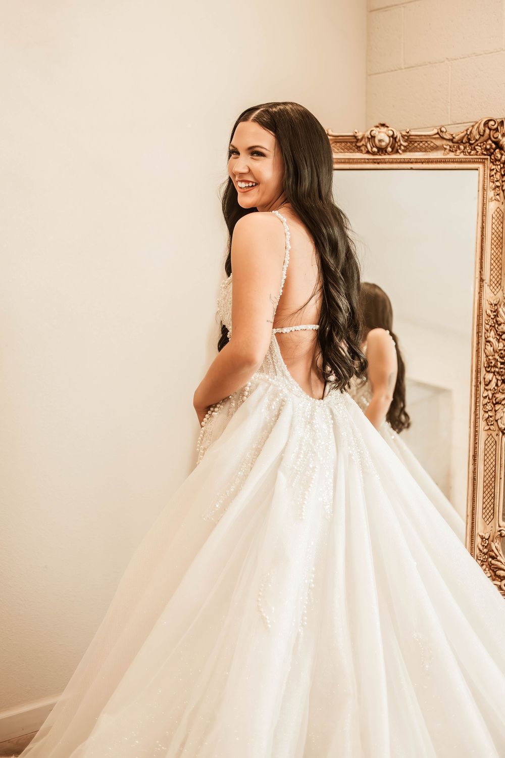 Bride in a stunning white ball gown with lace details and open back, smiling over her shoulder in front of a gold mirror.
