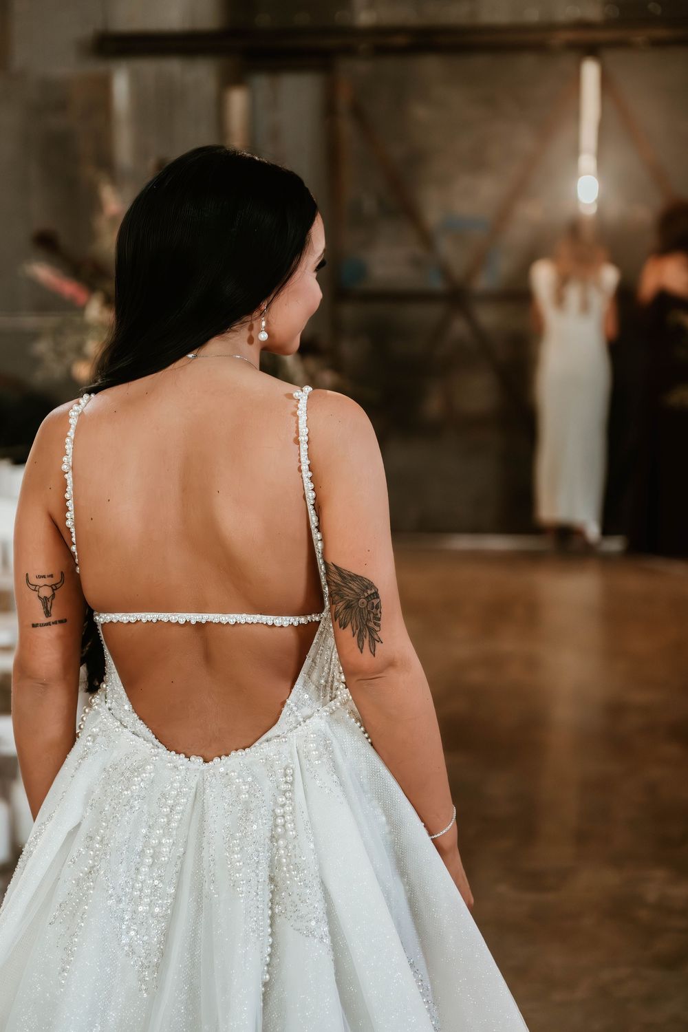 Bride in a backless beaded wedding gown with spaghetti straps and tattoos, viewed from behind at a reception.