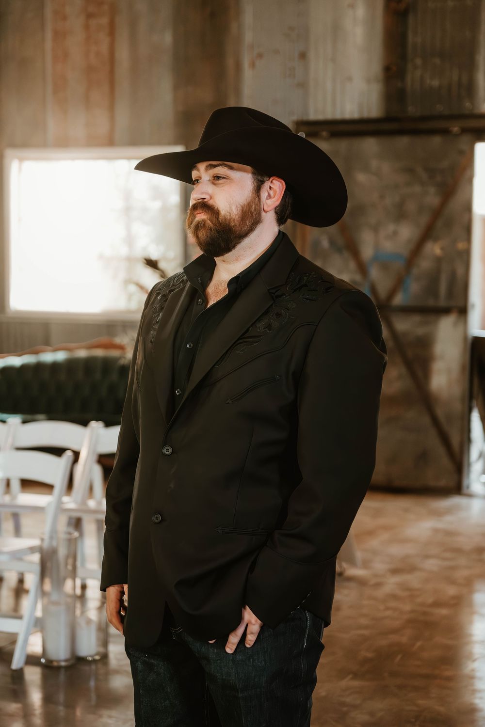 Bearded man in black cowboy hat and blazer stands indoors at a rustic venue with white chairs in background.