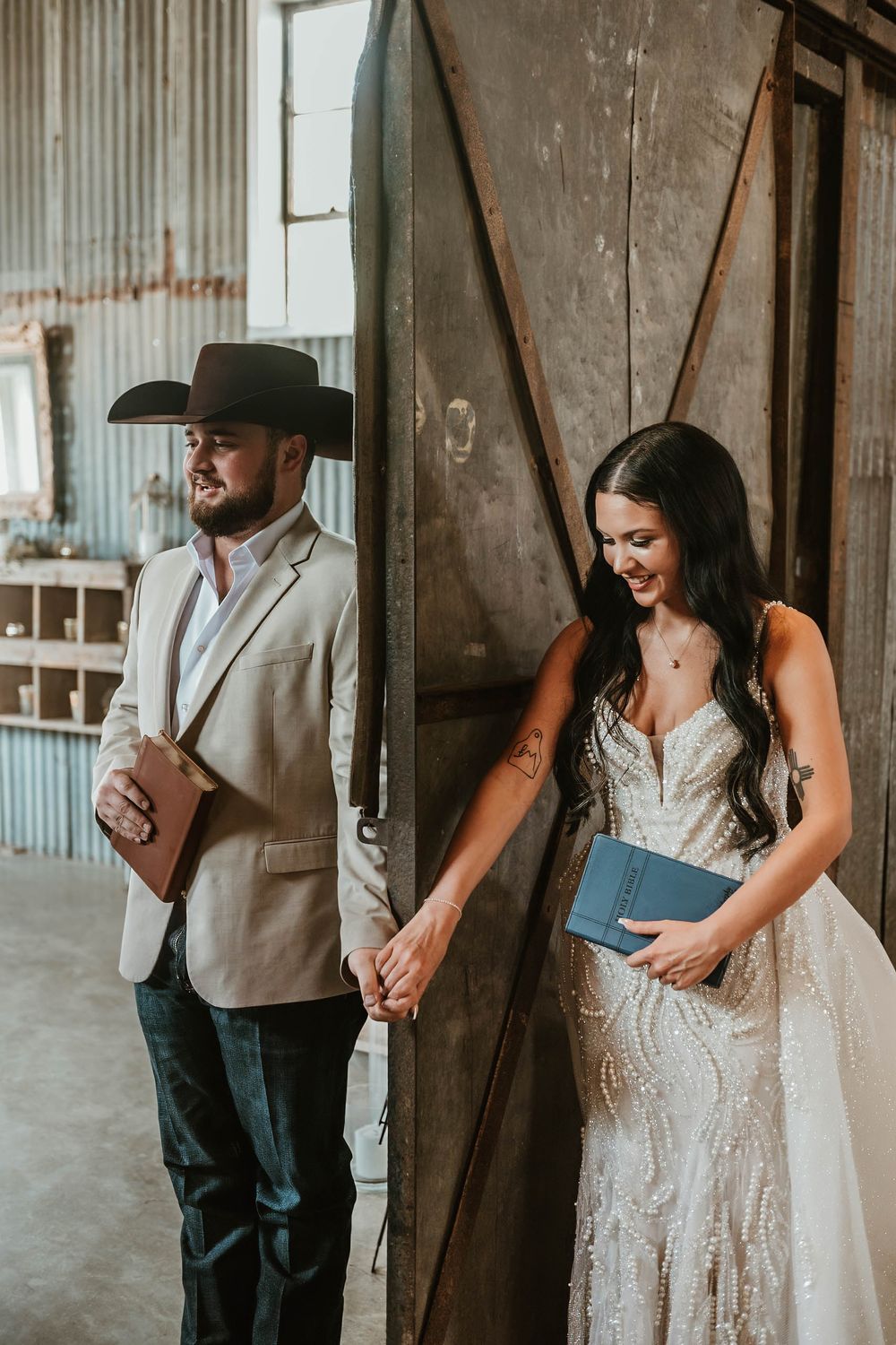 Bride in beaded gown and groom in tan suit with cowboy hat hold hands during rustic barn wedding first look.
