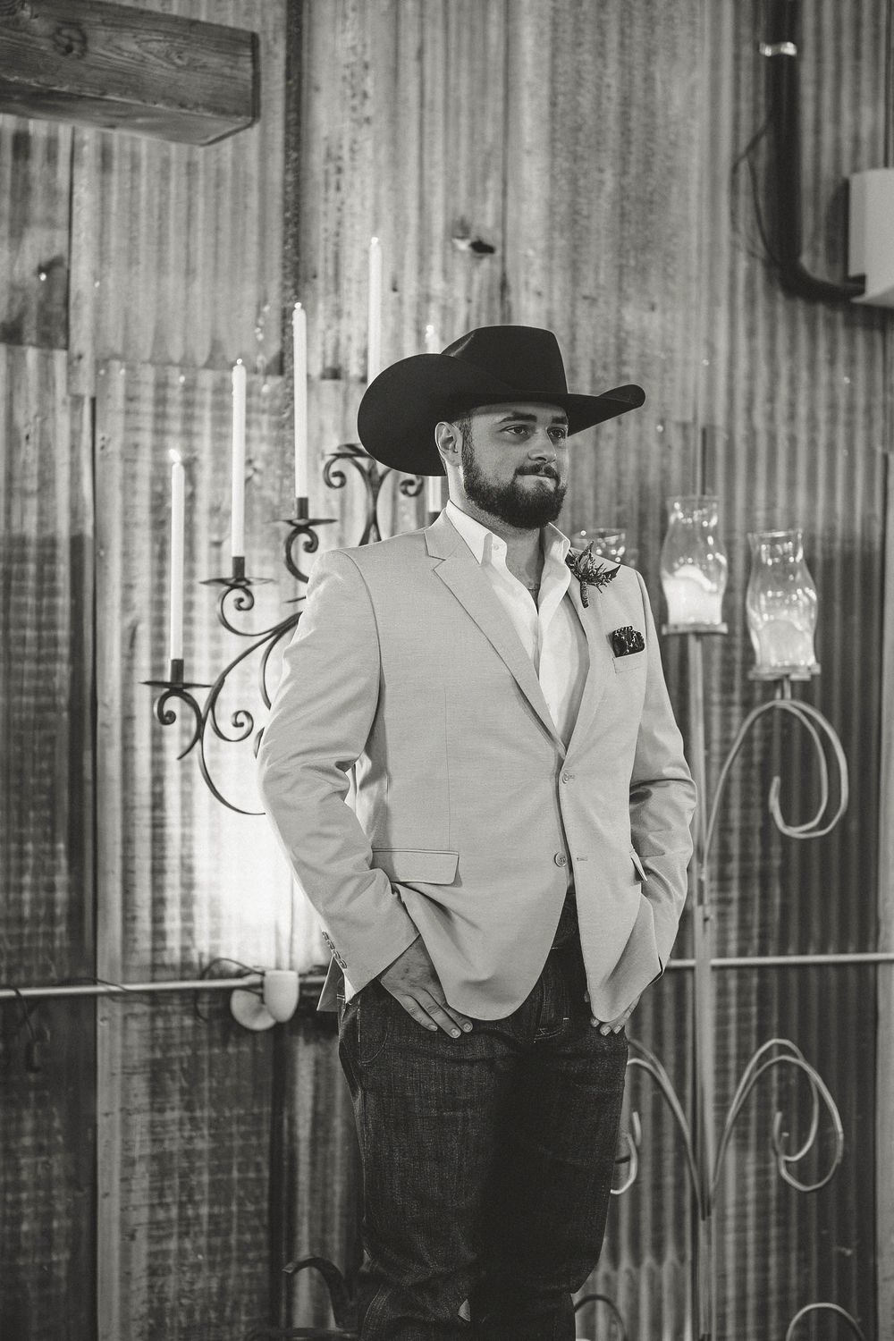 A bearded groom in a black cowboy hat and light blazer stands confidently in a rustic barn wedding venue.