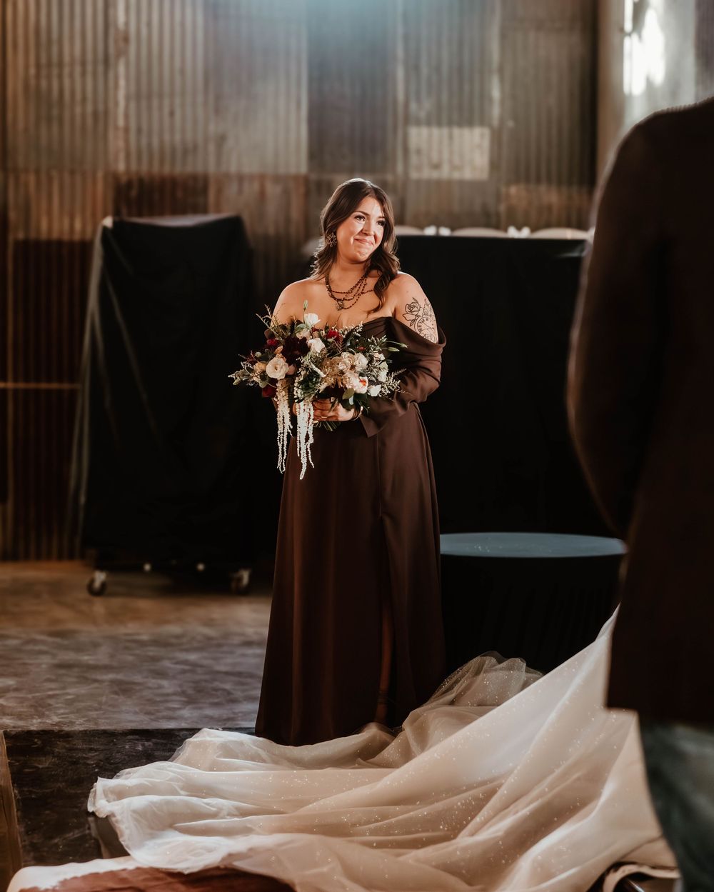 A smiling bridesmaid in a brown gown holds a bouquet inside a rustic barn venue.