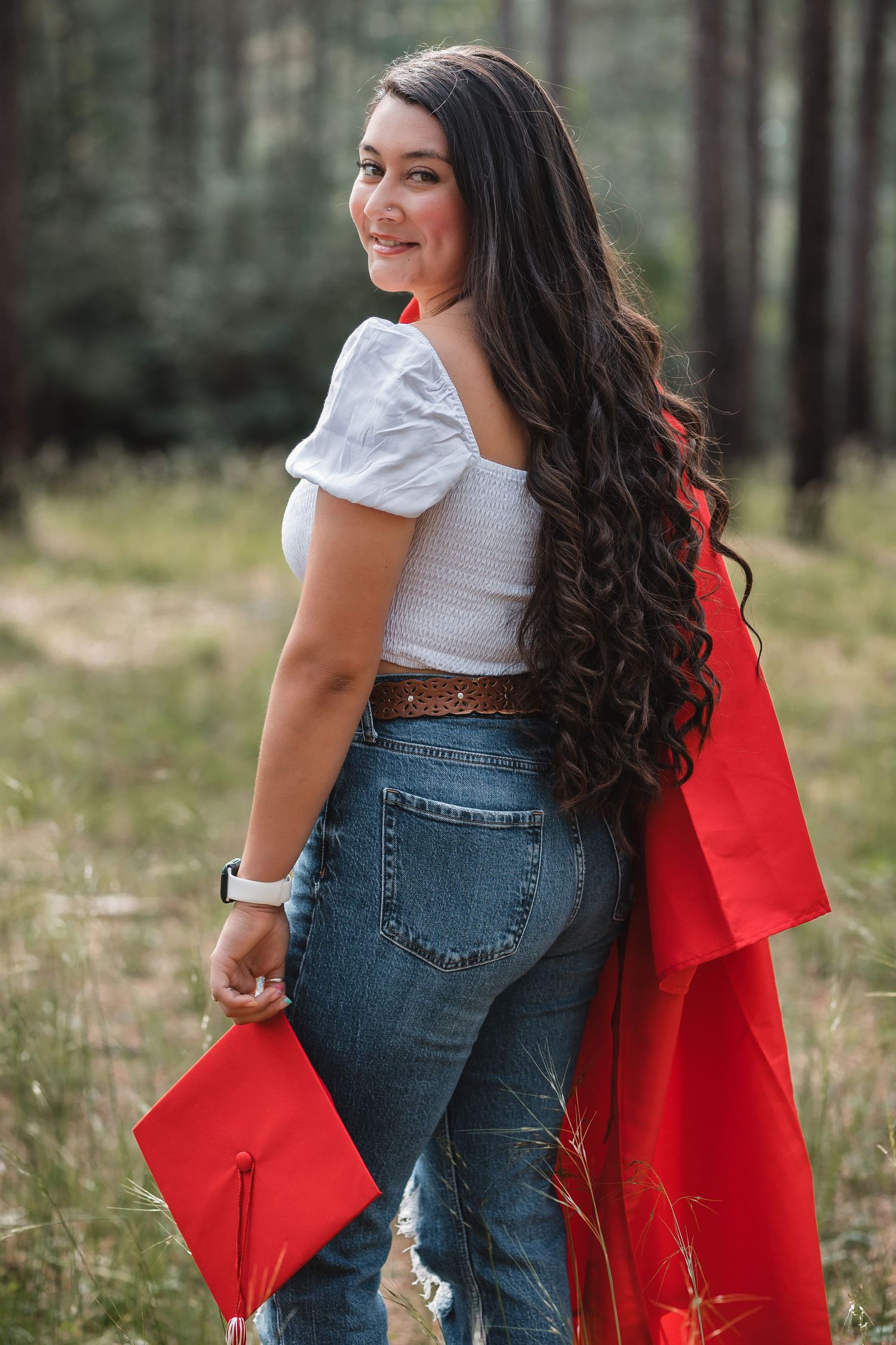 Smiling young woman in jeans and red graduation gown holding cap outdoors in forest setting.