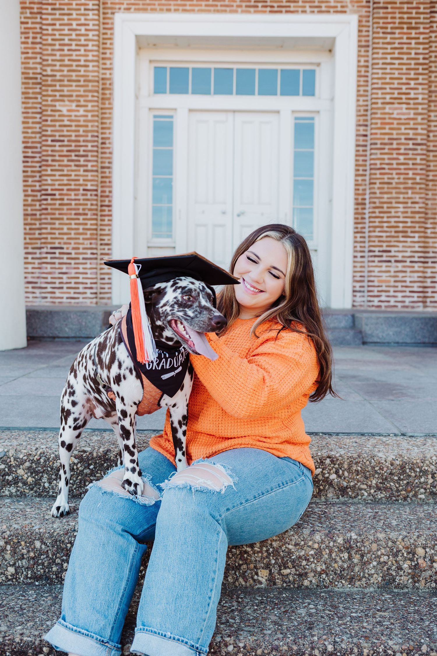 Young woman in orange sweater and jeans sits on steps with her Dalmatian dog wearing a graduation cap.