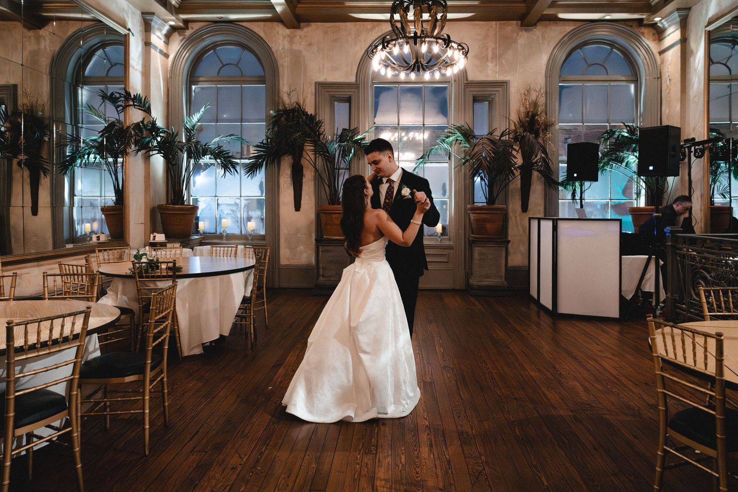 Bride and groom share first dance in elegant ballroom with hardwood floors, chandeliers, and gold chiavari chairs.