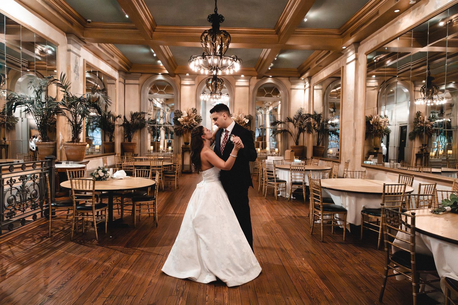 Bride and groom share first dance in elegant ballroom with chandeliers, mirrored walls, and gold chairs.