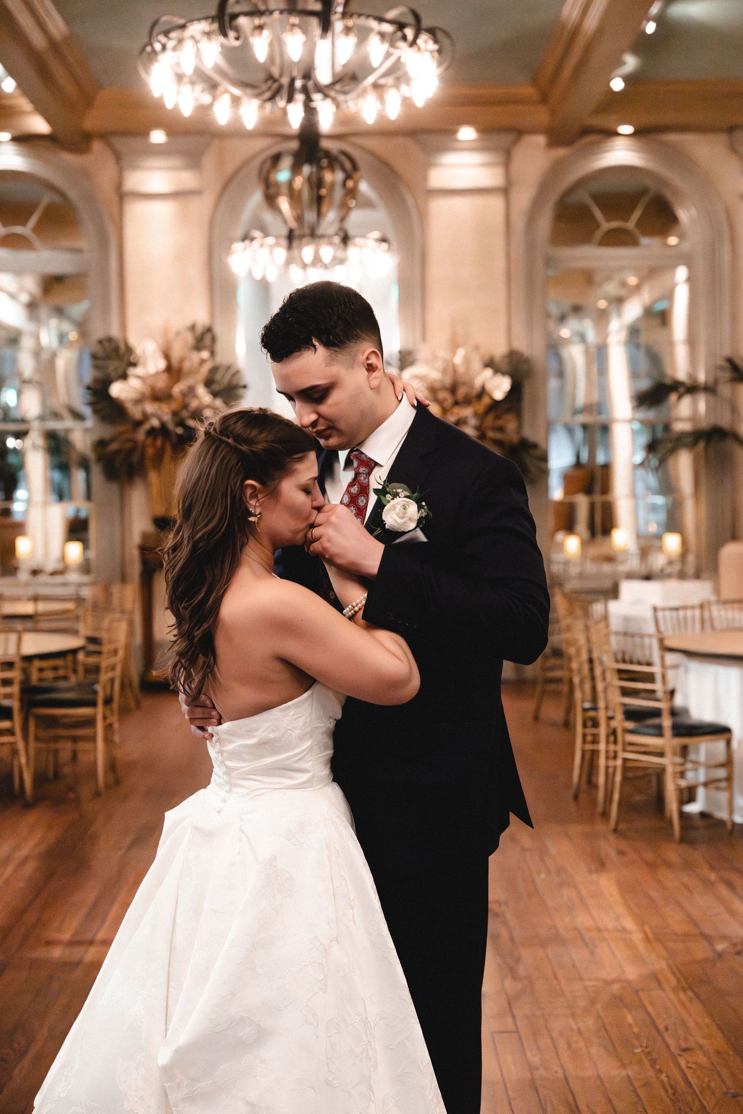 Bride and groom share first dance in elegant ballroom with chandelier and gold chiavari chairs at wedding reception.