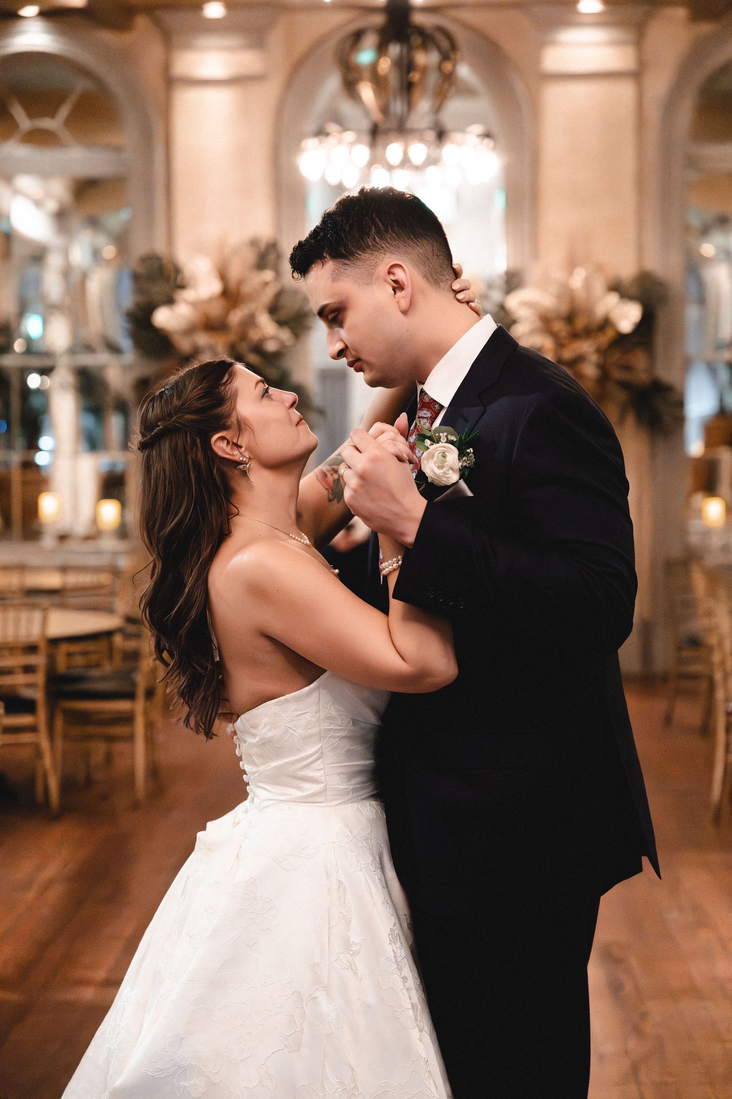 Bride and groom share their first dance in an elegant ballroom, gazing lovingly at each other in wedding attire.
