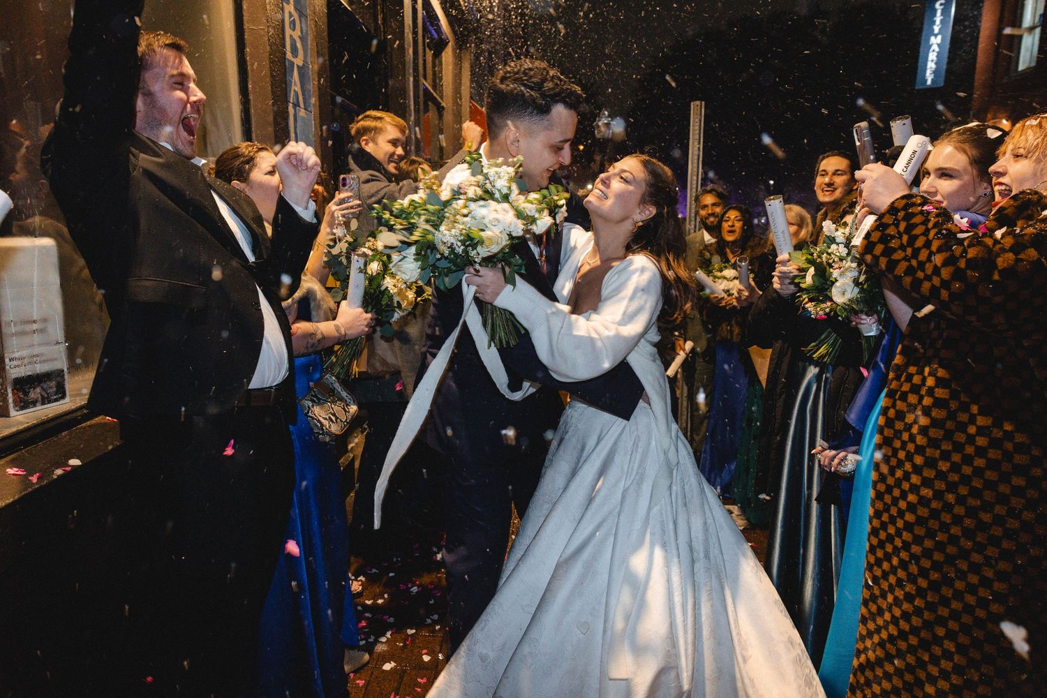 Bride and groom share a kiss surrounded by cheering guests throwing confetti outside a venue.