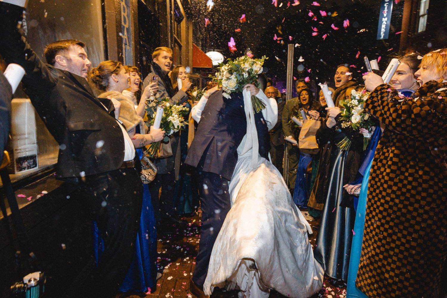 Bride and groom exit wedding venue through cheering guests throwing confetti and flower petals at night.