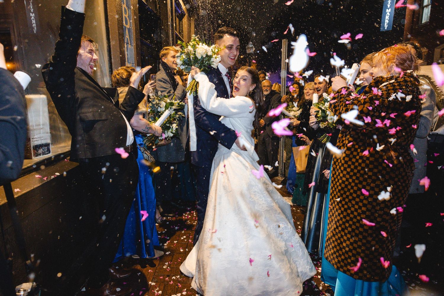 Bride and groom celebrate with confetti shower surrounded by guests at night wedding ceremony exit.