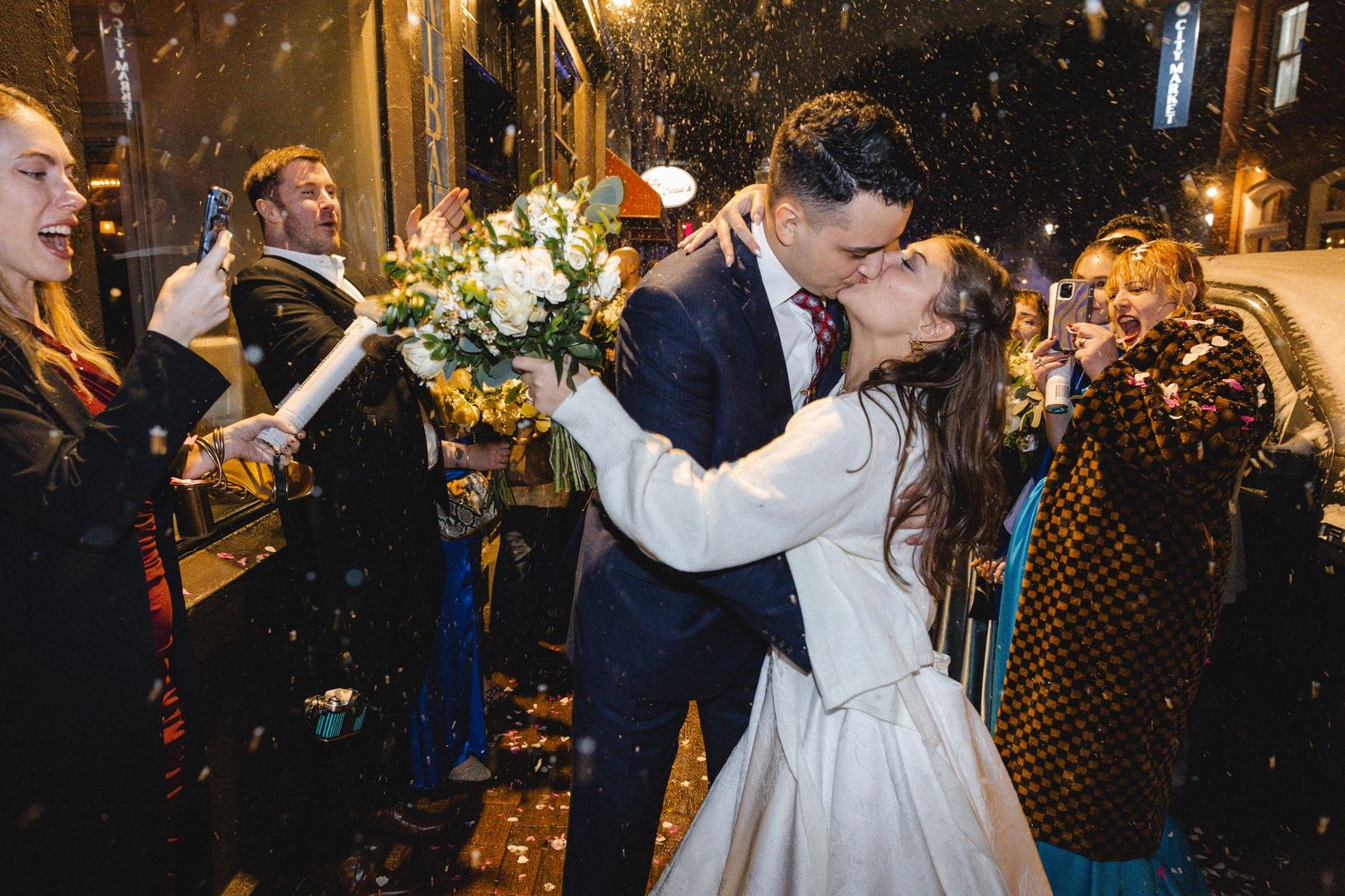 Newlywed couple sharing a kiss outdoors while guests celebrate with confetti and champagne at their wedding reception.