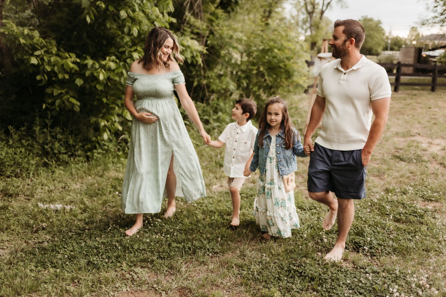 Pregnant woman in green dress walks with family outdoors, holding hands with two children and partner in sunny garden.