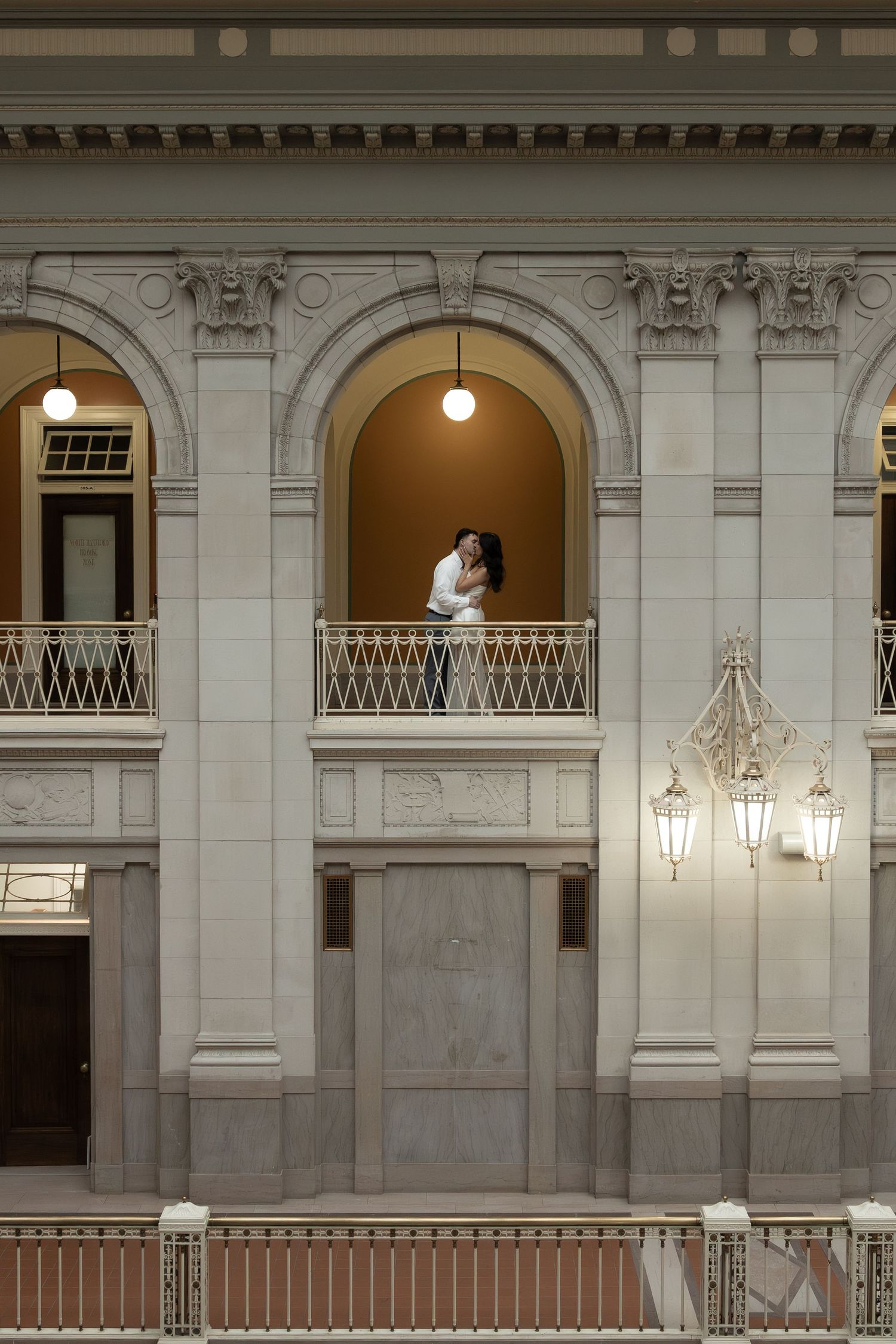 Couple embracing on an ornate balcony inside a grand classical building with arched windows and elegant stone columns.