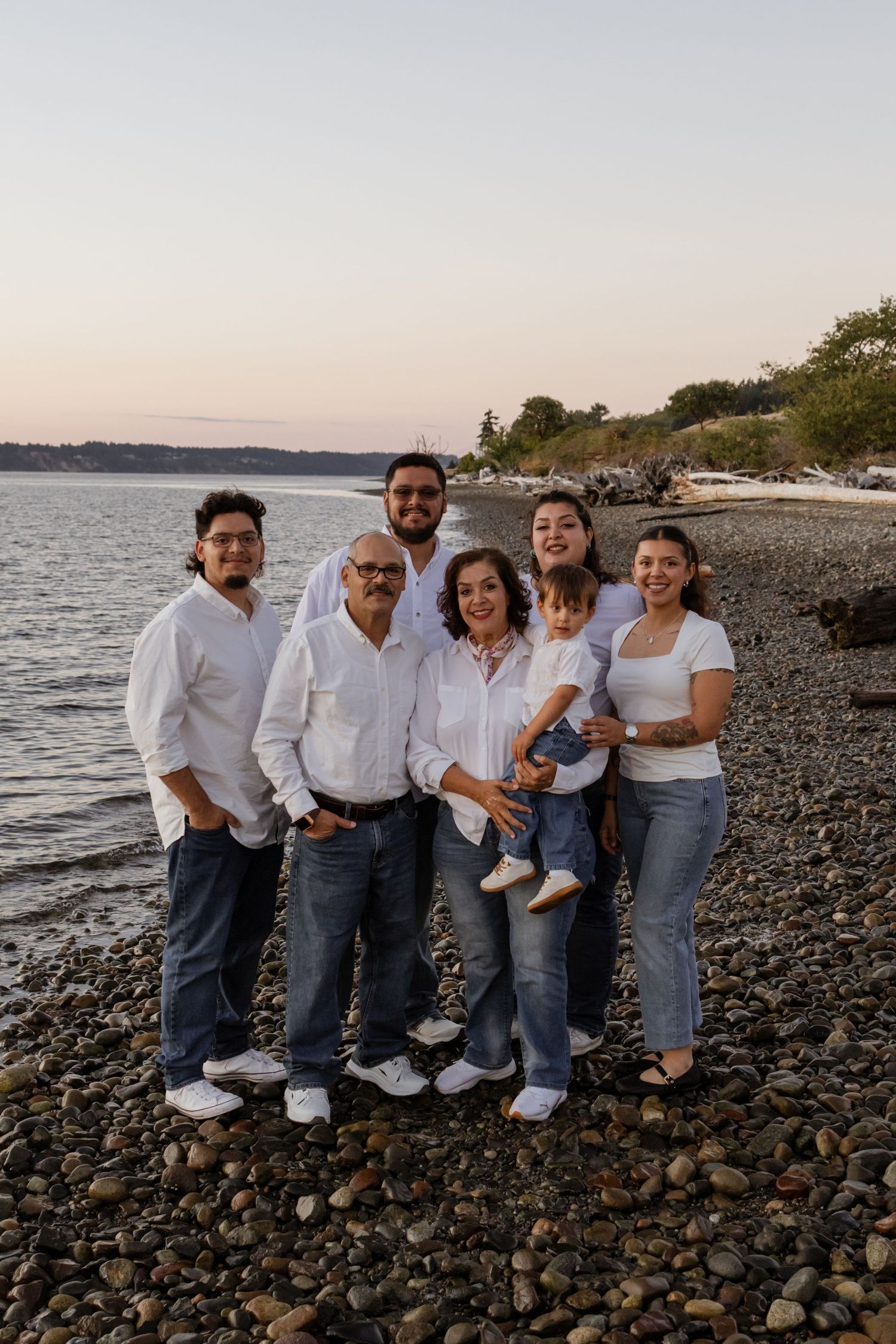 Family of seven posing together on a rocky beach at sunset, all wearing white tops and blue jeans.