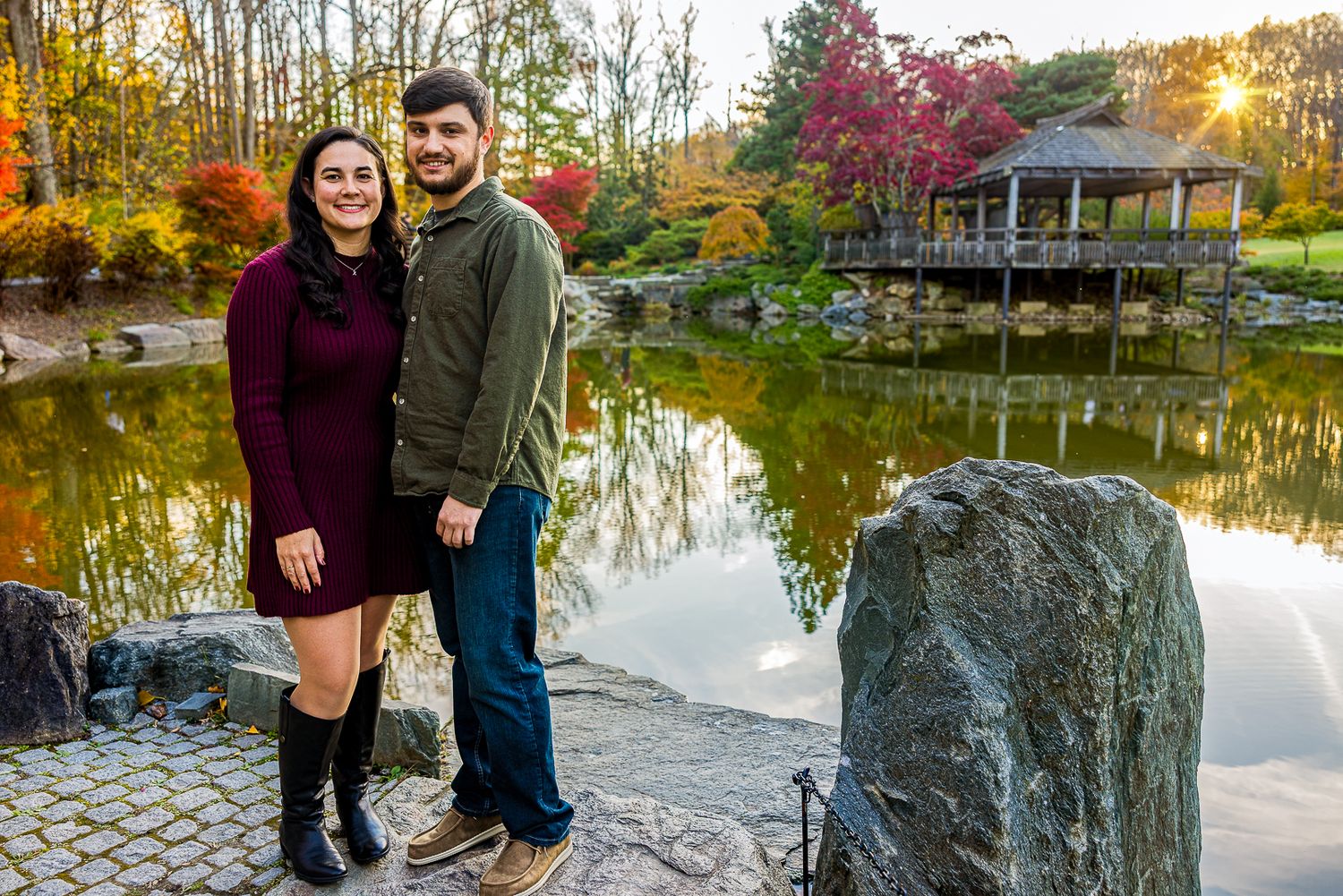 Couple posing by a serene autumn pond with colorful foliage and a gazebo in a Japanese garden.
