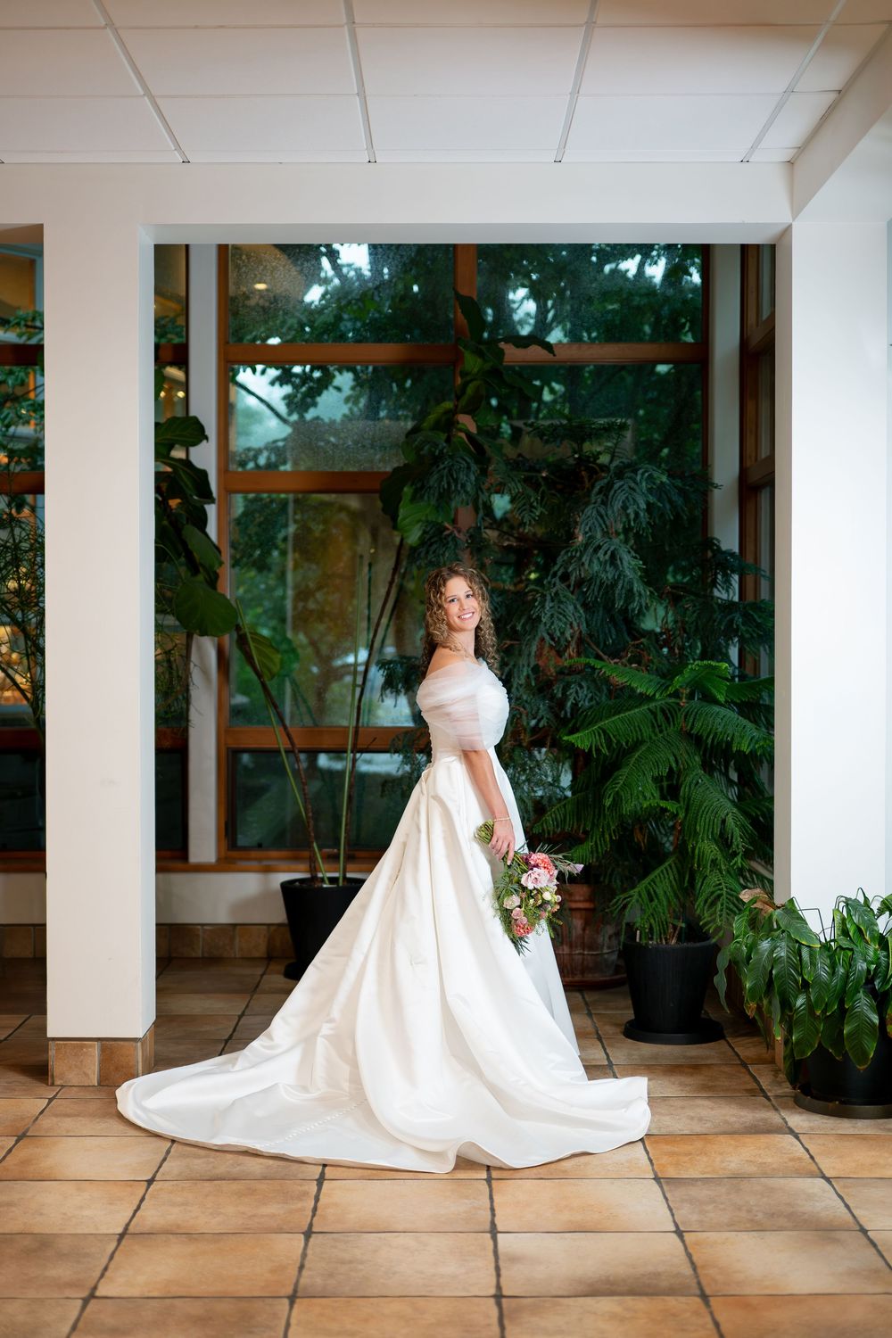 Elegant bride in a white satin gown with long train poses in a sunlit atrium with tropical plants.