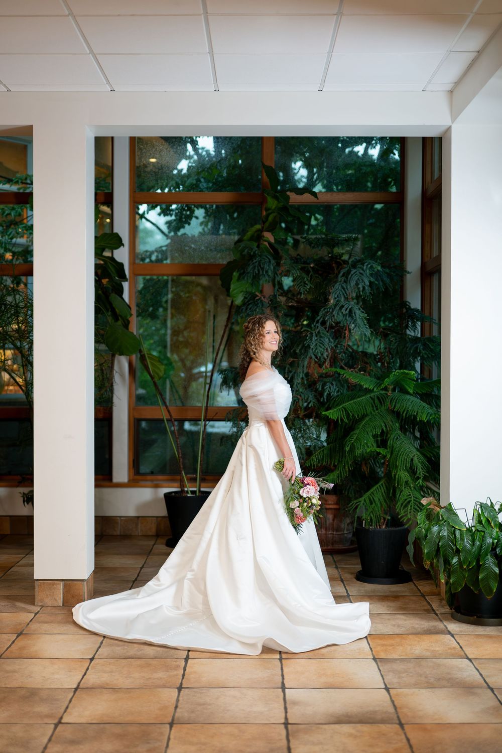 Bride in elegant white ball gown with long train holds bouquet in sunlit venue with lush green plants.