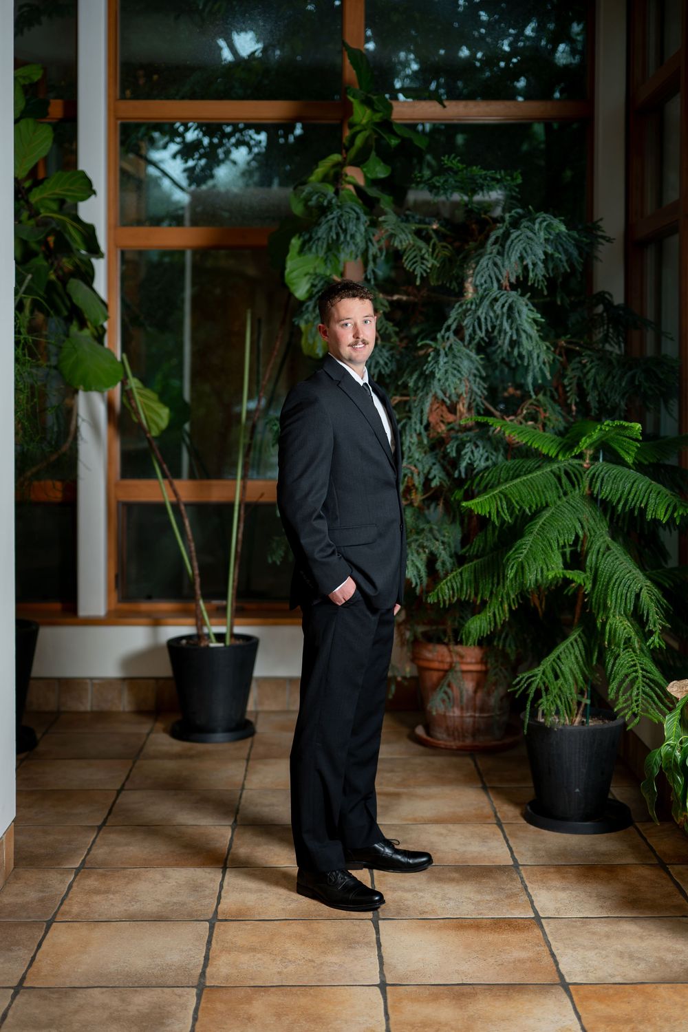 Dapper groom in a black tuxedo stands confidently in a plant-filled indoor venue with large windows.
