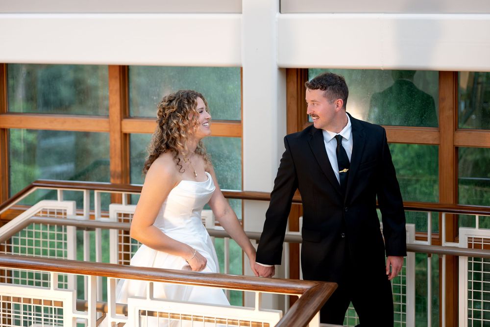 Bride and groom holding hands on an indoor balcony, smiling at each other in wedding attire.