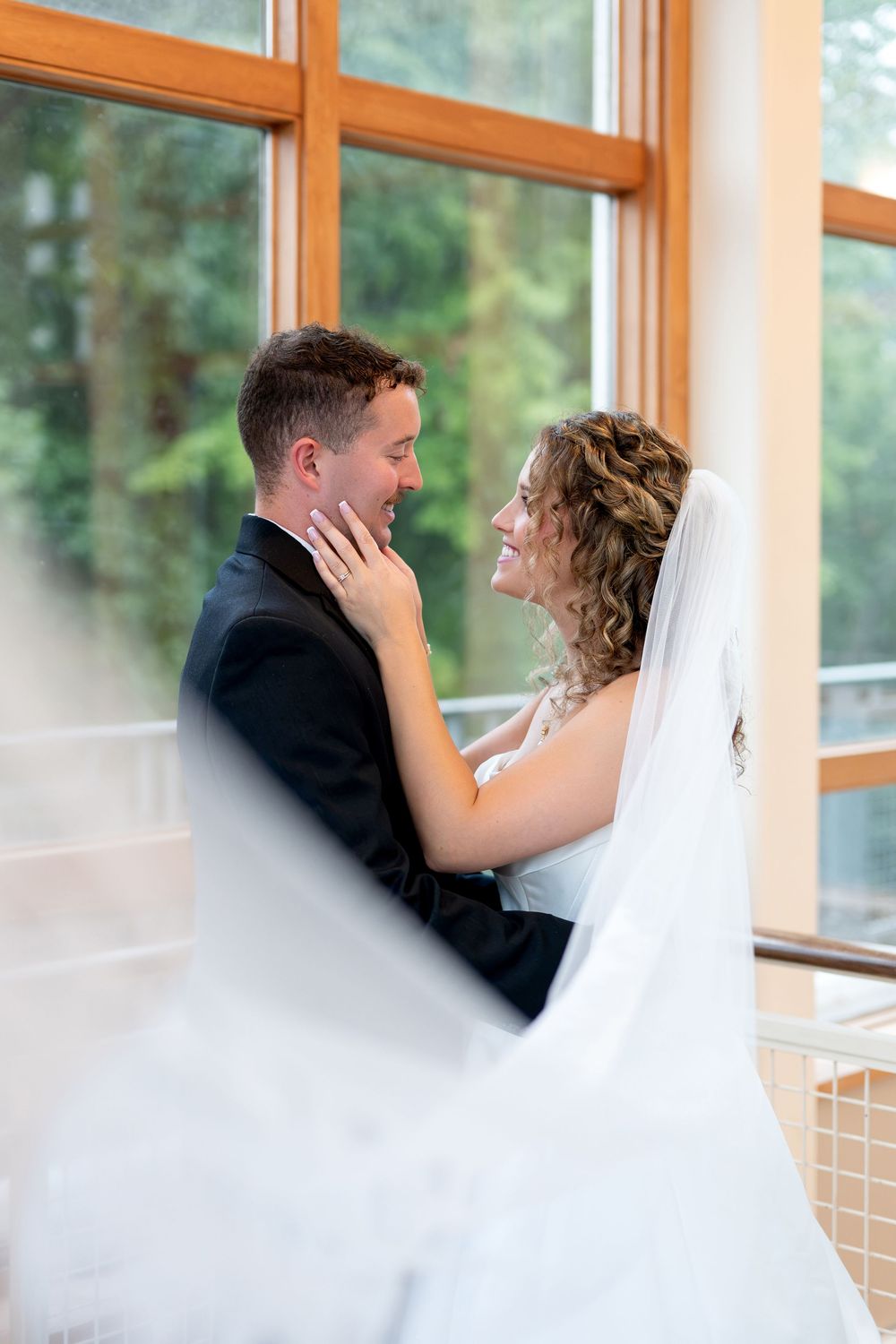 A bride and groom share a romantic moment indoors, the bride in a white veil gently touching the grooms face.