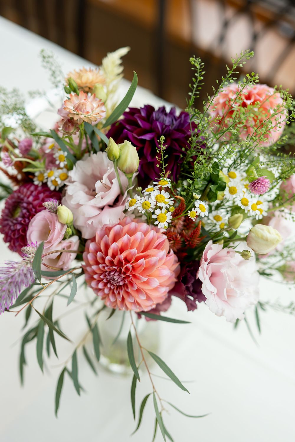Elegant floral arrangement with pink dahlias, purple flowers, daisies, and greenery in a glass vase on a white table.