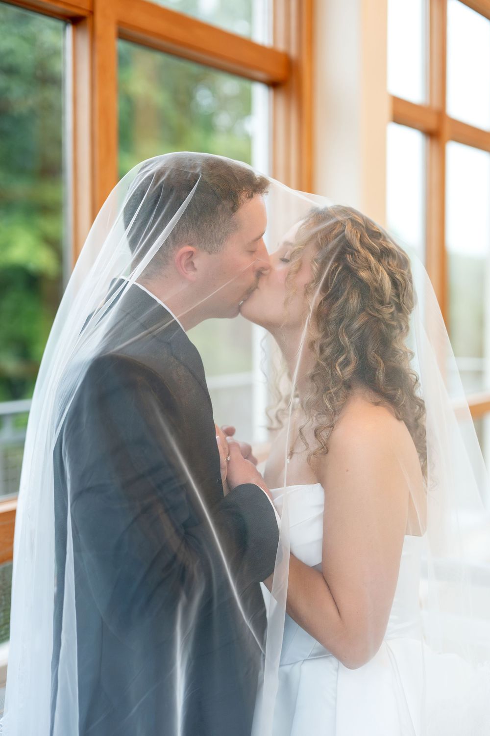 Bride and groom share a tender kiss under a flowing wedding veil near bright windows.