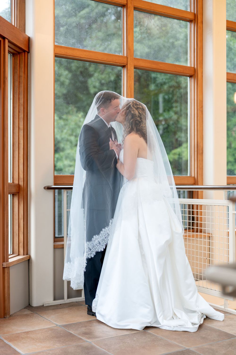 Bride and groom sharing a romantic kiss under a wedding veil by large windows in an elegant indoor venue.