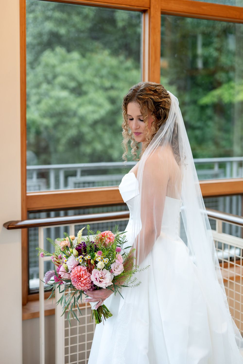 Elegant bride in a white strapless gown holding a colorful bouquet while standing near large wooden-framed windows.