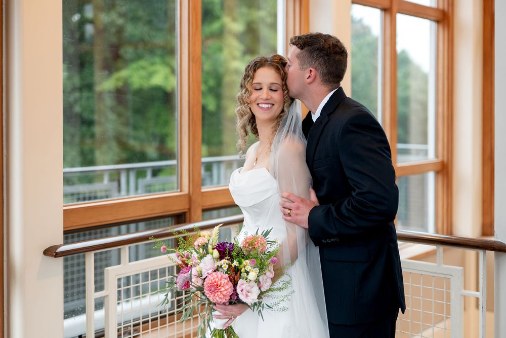 Groom in black suit kissing bride on cheek as she smiles, holding a vibrant floral bouquet indoors.