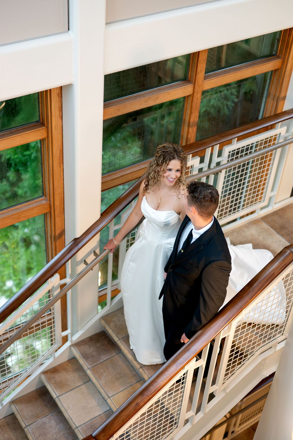 Bride and groom hold hands on a staircase, smiling at each other in a bright modern venue.