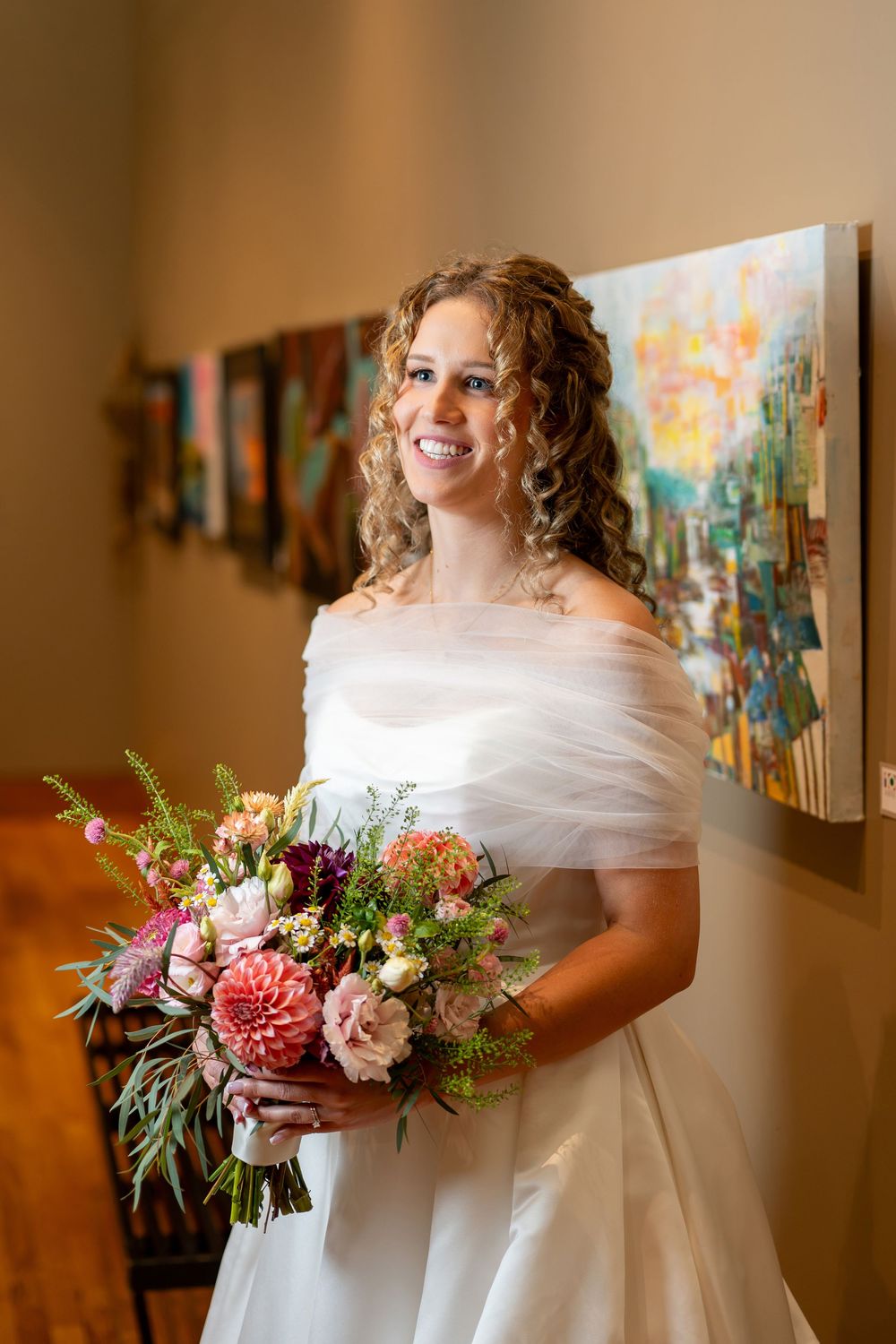 Smiling bride in off-shoulder white gown holds colorful bouquet with dahlias and roses in front of art gallery wall.