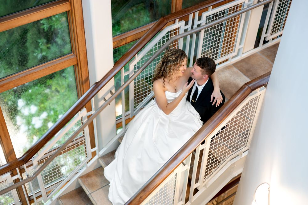 Bride in white ball gown and groom in black suit share a tender moment on elegant staircase.