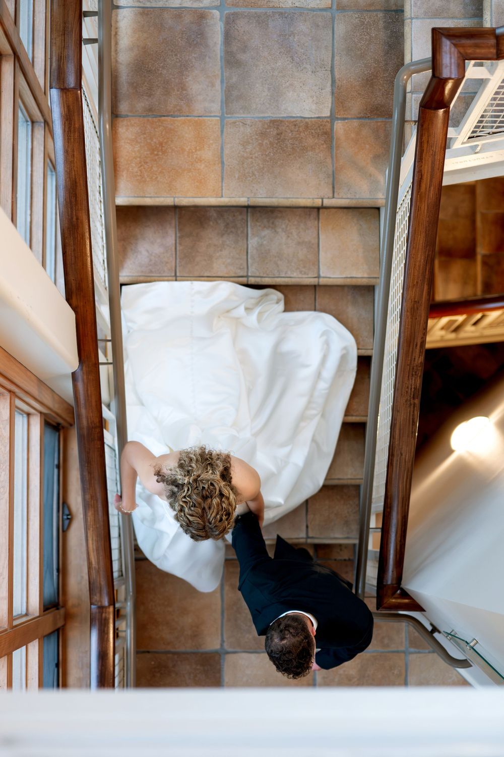 Aerial view of bride in white gown and groom in black suit descending tiled staircase together.
